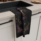 Vespertine Gothic Moon Bat microfiber tea towel draped beside a kitchen sink, showing black fabric with purple and gold celestial artwork